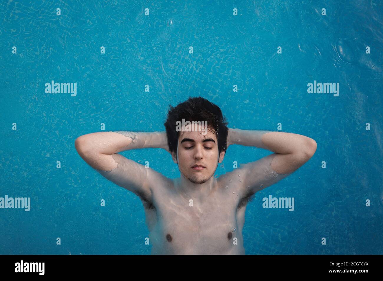 Draufsicht auf einen jungen Mann mit geschlossenen Augen, der in einem Schwimmbad mit den Händen im Hinterkopf schwimmt. Genießen Sie Urlaub beim Sonnenbaden Stockfoto