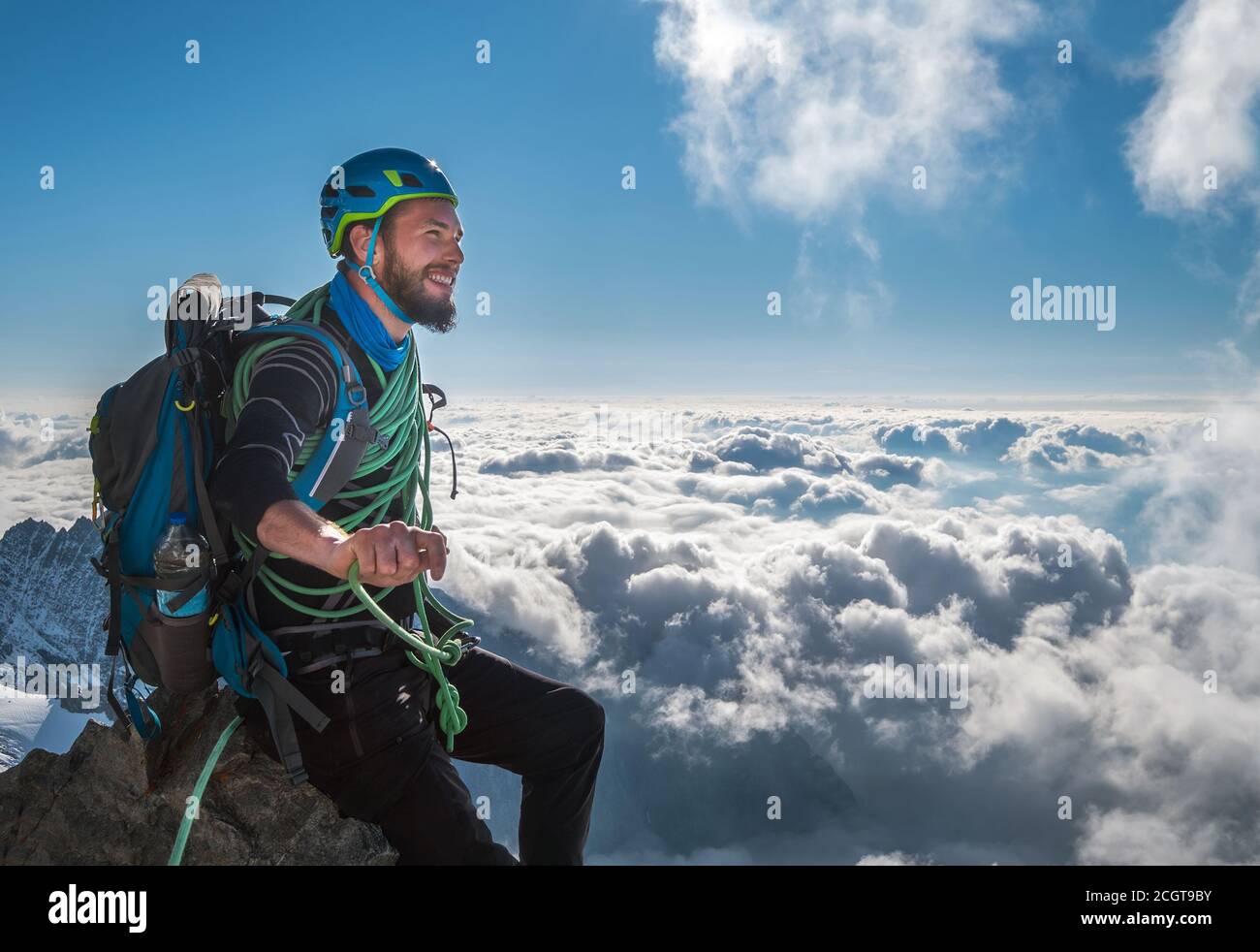 Lächelnder bärtiger Kletterer in einem Sicherheitsgeschirr, Helm und am Körper gewickeltem Kletterseil, der auf 3600m Höhe auf einer Klippe sitzt und malerisch anschaut Stockfoto