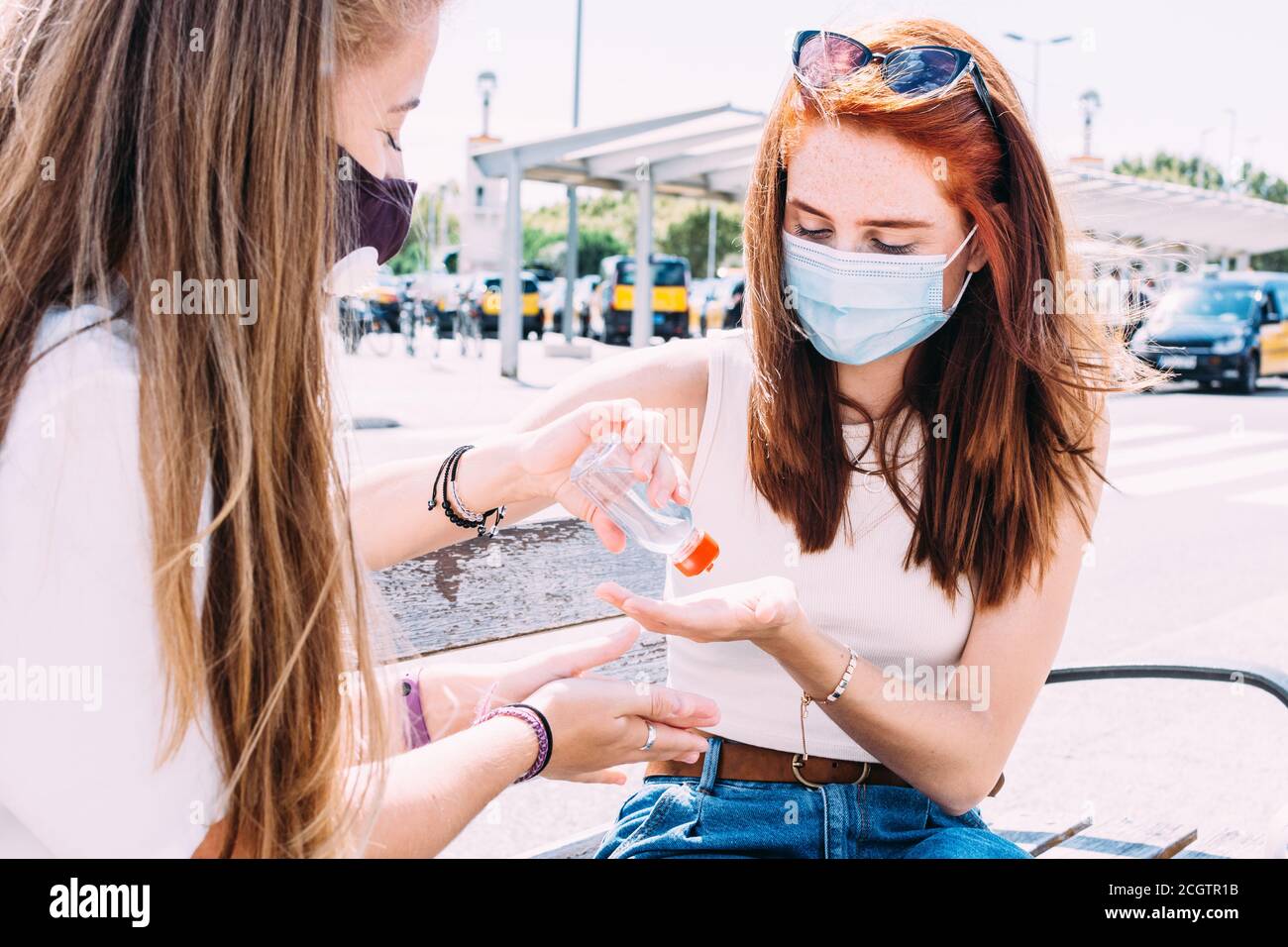 Zwei junge Frauen sitzen auf einer Bank vor dem Zug Station auf antibakterielles Gel setzen Stockfoto