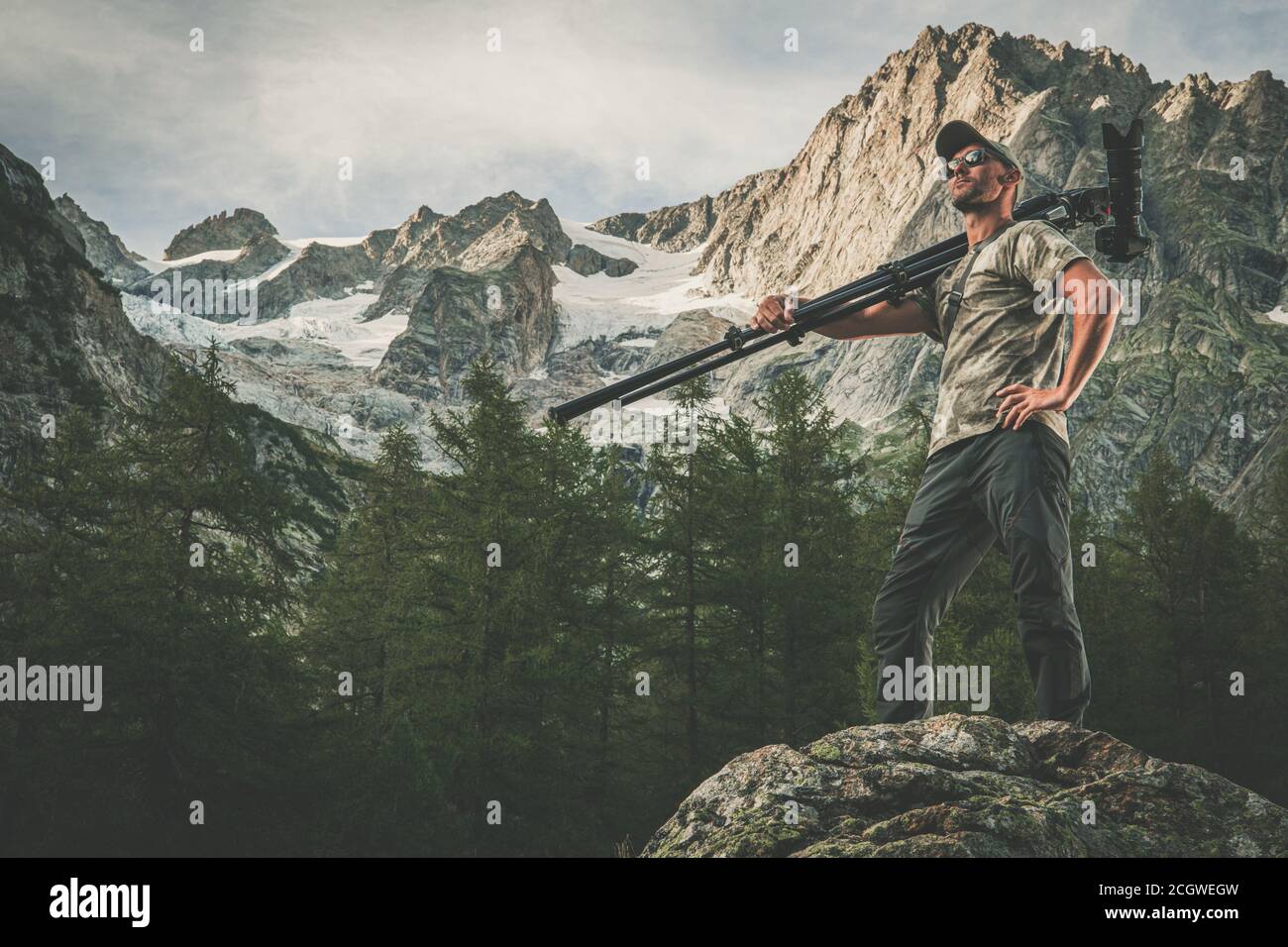 Kaukasischer kommerzieller Naturfotograf in den 40er Jahren mit professioneller Fotoausrüstung auf der Schulter. Digitalkamera auf einem Stativ. Alpine Landschaft in Stockfoto