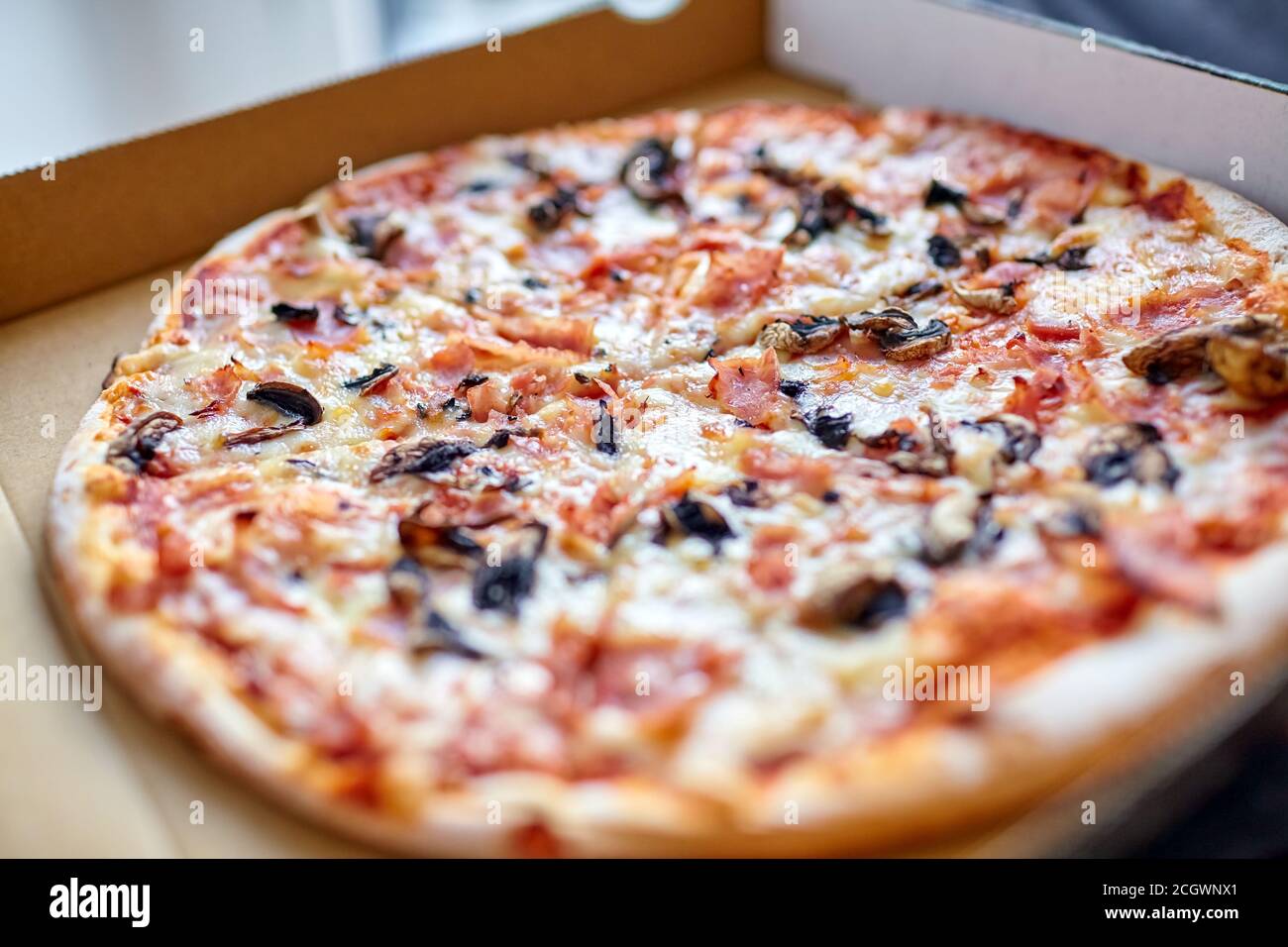 Ganze Pizza mit Pilzen, Käse und Schinken in einer Kartonschachtel. Nahaufnahme. Selektiver Fokus Stockfoto