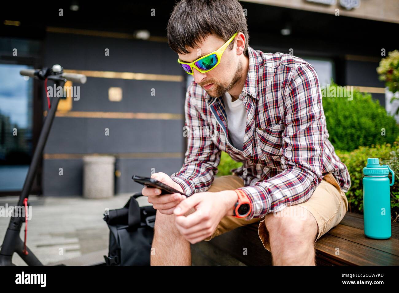 Kaukasischen Mann nutzt moderne Technologien Internet und Mobilfunk auf Smartphone, während auf der Bank in der Stadt in der Nähe Elektroroller sitzen. Ökologie Stockfoto