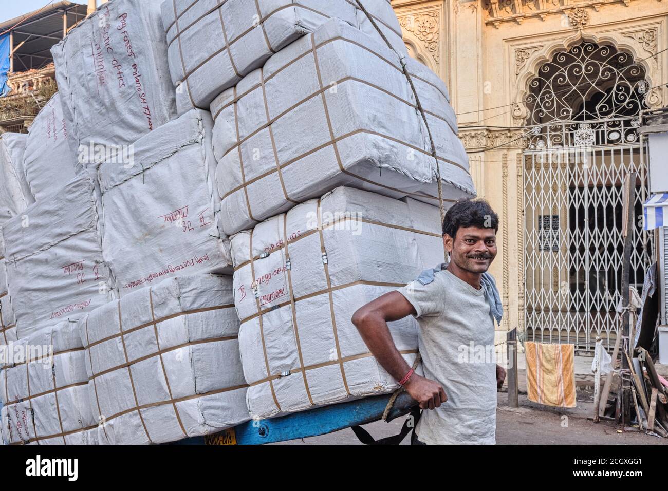 Ein fröhlicher Handkarrenabzieher in Mumbai, Indien, zieht einen Wagen, der mit fast einer Tonne Ballen Stoff beladen ist Stockfoto