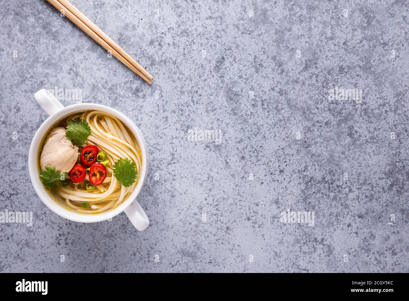 Asiatische Hühnersuppe. Bouillon und Fleisch, Chili, Koriander, Frühlingszwiebeln, japanische Nudeln in weißer Schale auf grauer Oberfläche. Essstäbchen aus Holz in der Nähe des Behälters Stockfoto