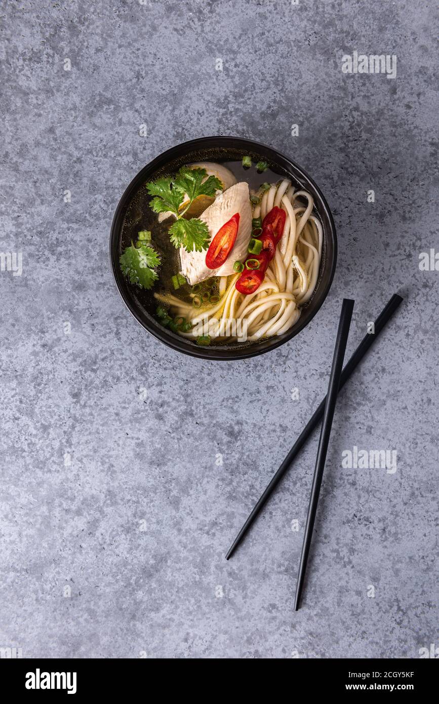 Würzige asiatische Suppe. Hühnerbouillon und Fleisch, rote Chilischote, Koriander, Frühlingszwiebeln und japanische Nudeln in schwarzer Suppenschüssel mit Essstäbchen auf grauem Su Stockfoto