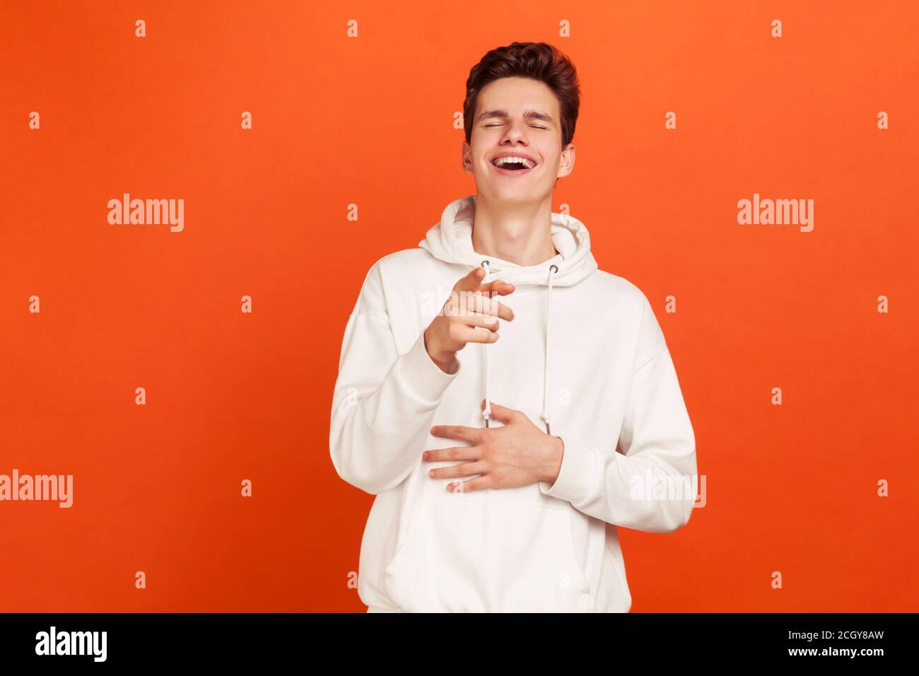 Glücklich zufrieden junger Mann in weißen Hoodie zeigt Finger auf Sie und aufrichtig lachen Hand auf Bauch halten. Spaß haben. Innenaufnahme des Studios isoliert Stockfoto