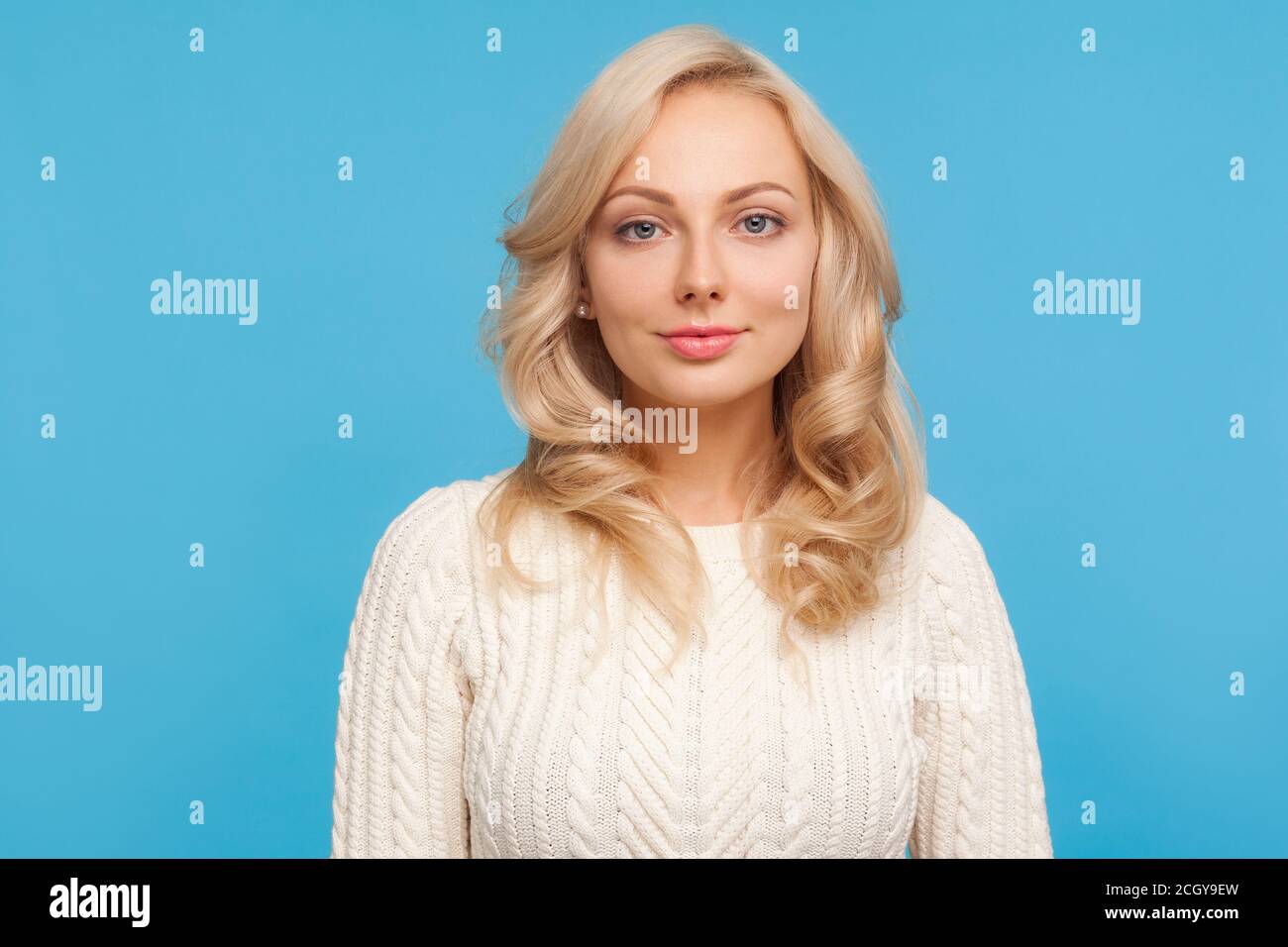 Porträt von schönen selbstbewussten Frau mit lockigen blonden Haaren Blick auf die Kamera mit leichtem Lächeln auf Gesicht. Innenaufnahme des Studios isoliert auf blauem Hintergrund Stockfoto
