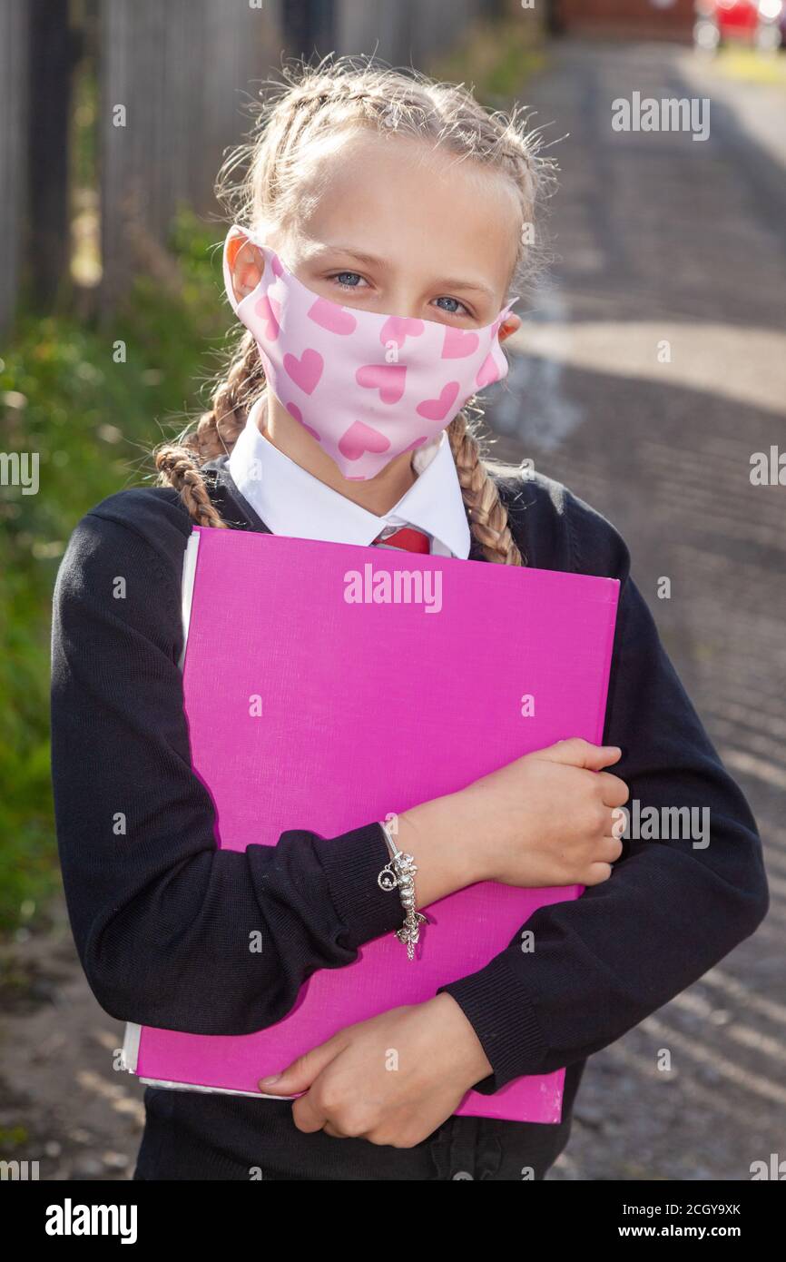 Eine zehnjährige Schülerin mit ihren Haaren in Zöpfen, die einen pinken A4-Ordner in der Hand halten und eine Gesichtsmaske tragen. Stockfoto