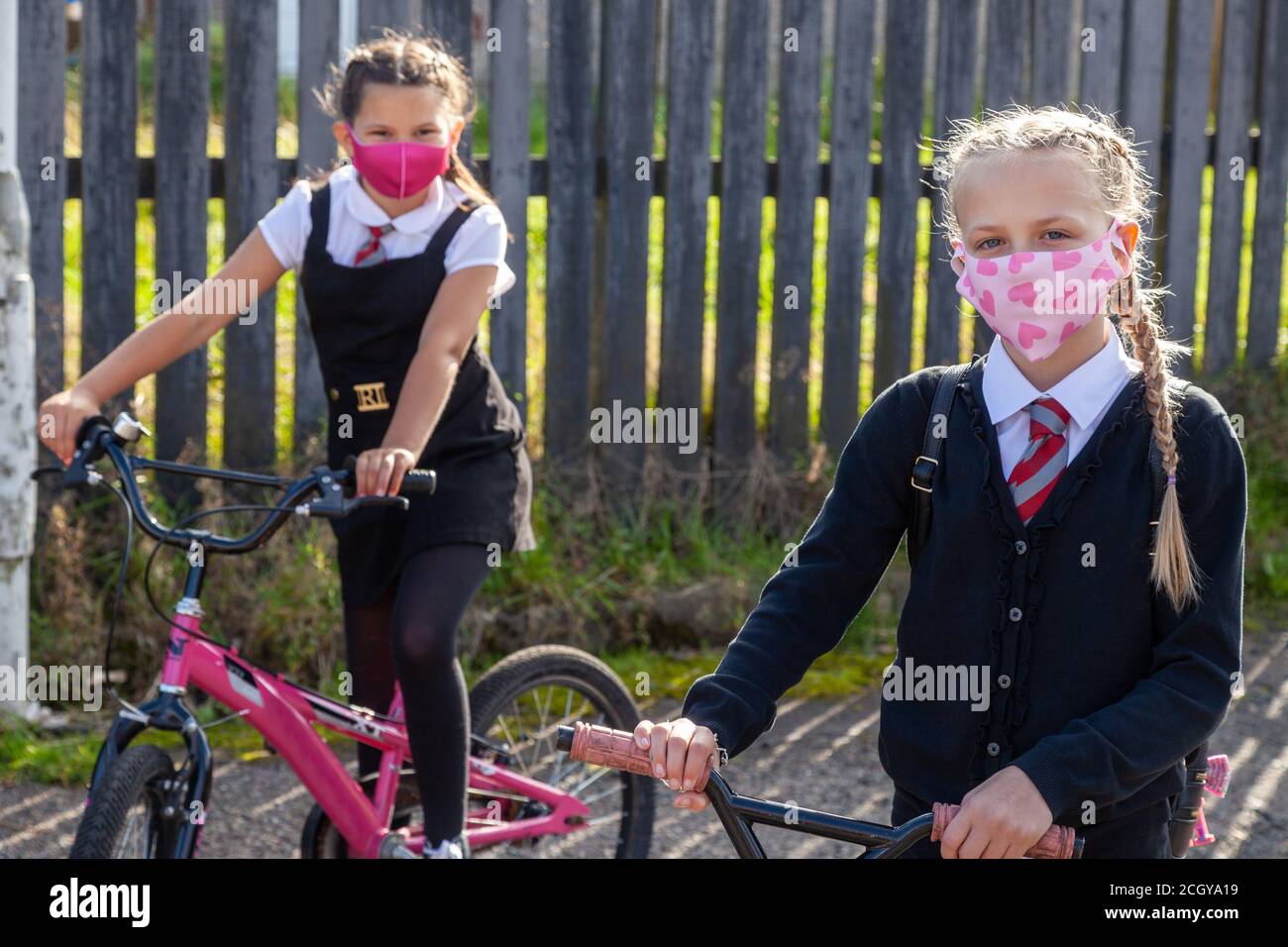 Zwei zehnjährige Schulfreunde in Schuluniformen sitzen auf Fahrrädern und tragen Gesichtsmasken. Stockfoto