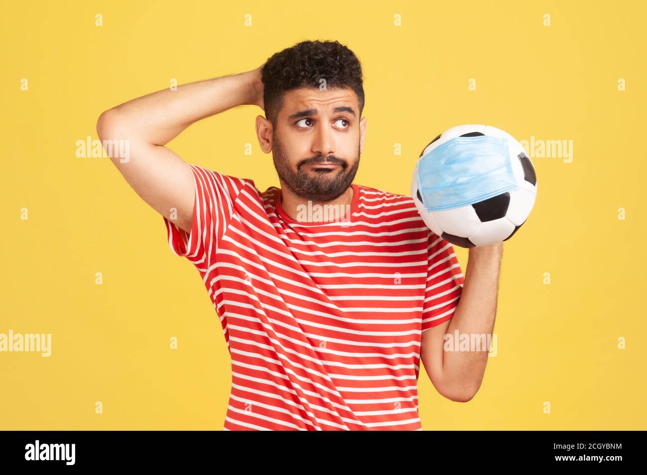 Trauriger nachdenklicher Mann im roten T-Shirt, der den Fußball in Schutzmaske hält, Symbol des Fußballwettbewerbs während des covid-19 Virus, Score-Fälschung. ICH Stockfoto