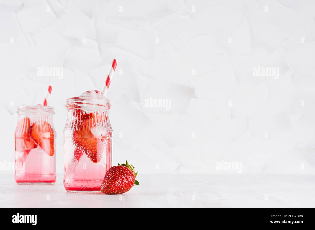 Erfrischende kalte Fruchtgetränke mit roter Erdbeere, Eis und Stroh in zwei transparenten Gläsern im eleganten weißen Interieur auf Holztisch, Kopierraum. Stockfoto