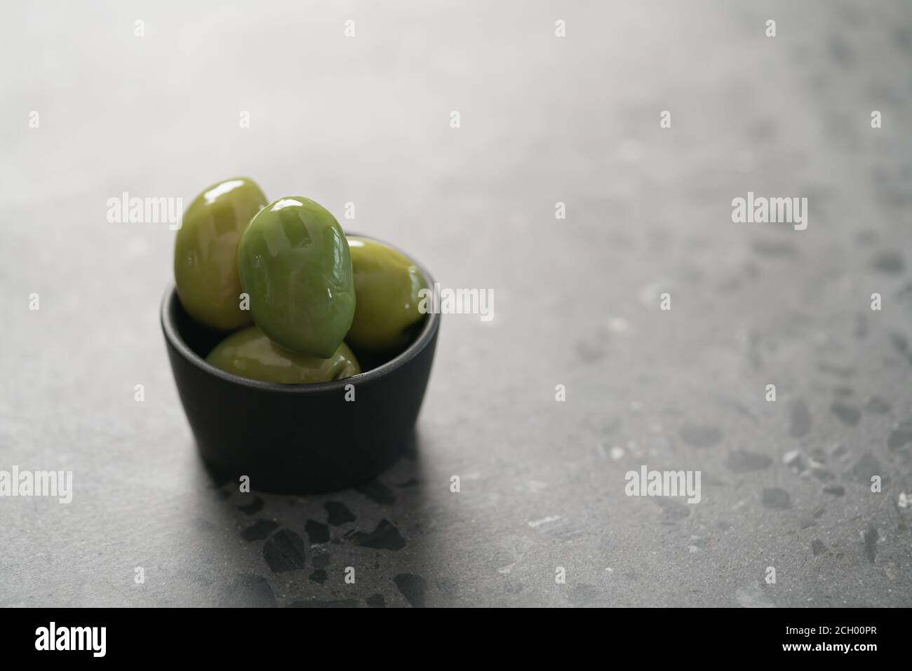 Große grüne Oliven in schwarzer Schale auf Beton Hintergrund, flacher Fokus Stockfoto