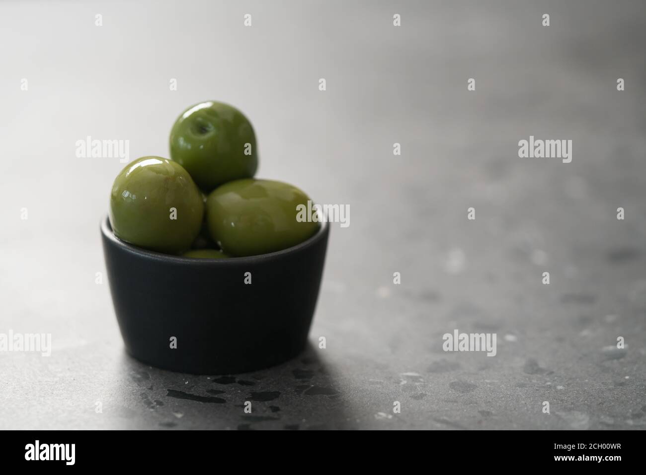 Große grüne Oliven in schwarzer Schale auf Beton Hintergrund, flacher Fokus Stockfoto