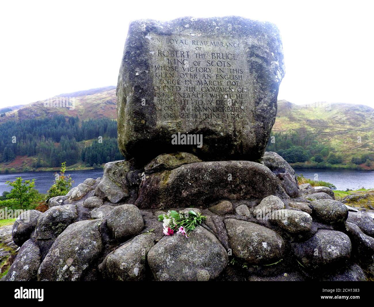Ein Blumenschmuck am Robert the Bruce (Earl of Carrick) Memorial, nahe Loch Troon, Galloway (2020 Foto). - die Schlacht von Glen Trool fand im ersten Krieg der schottischen Unabhängigkeit statt, im April 1307. Glen Trool ist ein schmaler glen im südlichen Hochland von Galloway, Schottland, in dem sich Loch Trool (hinter dem Denkmal) befindet. Als sich die englischen Soldaten in einer einzigen Datei dem schmalen glen näherten, schleuderten Bruce's Männer Felsbrocken und Pfeile nach unten auf sie. Die englischen Soldaten wurden getötet oder zogen sich zurück und Bruce siegte. Stockfoto