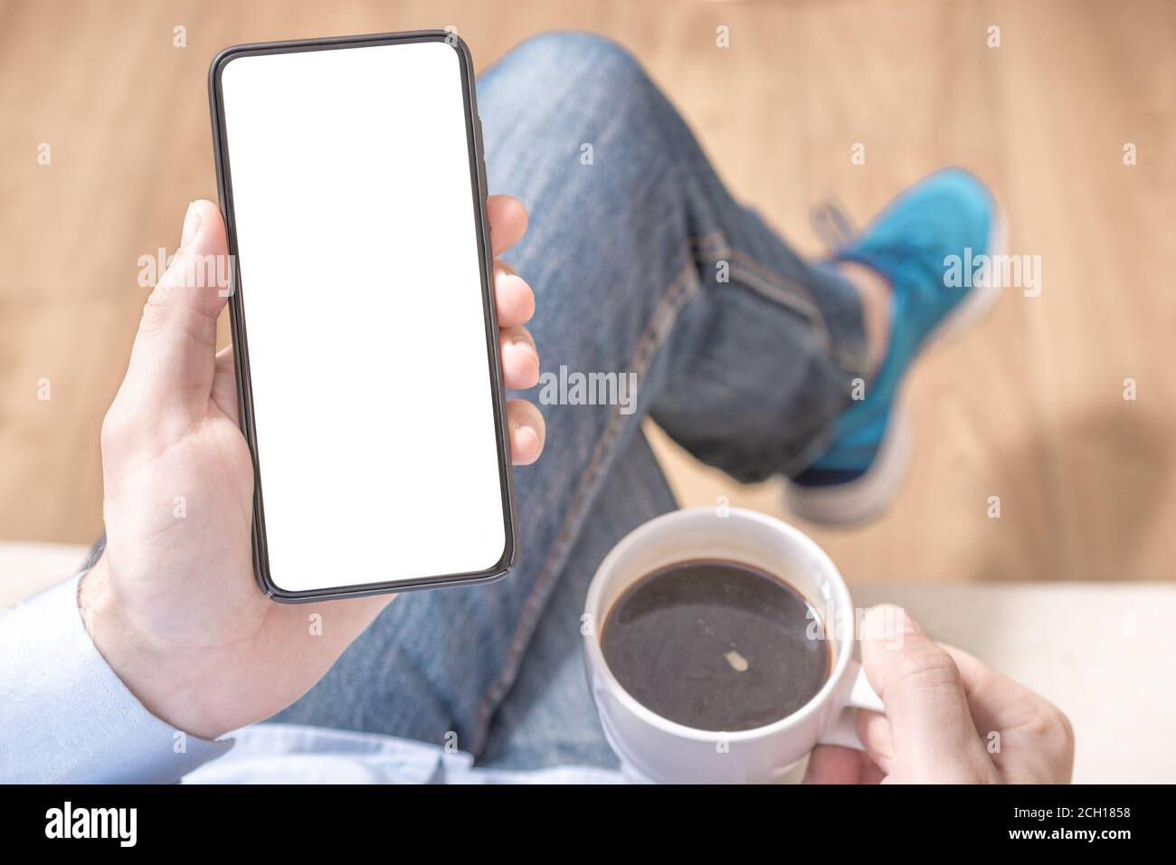 Handymockup. Mockup Bild der Hand hält schwarzes Handy mit leeren weißen Bildschirm auf Oberschenkel mit blauen Turnschuhen Schuhe an Vintage Fliese Boden in Stockfoto