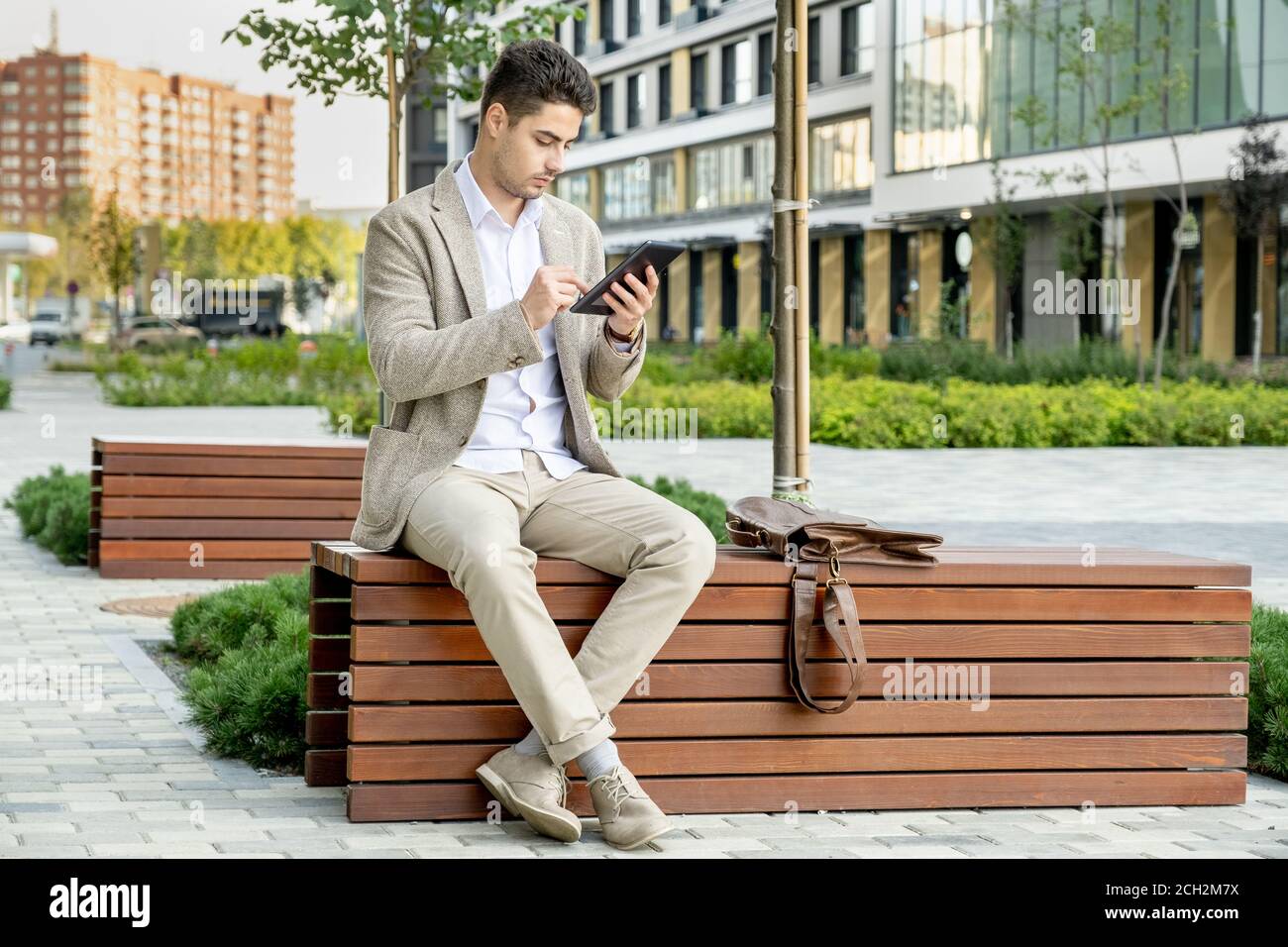 Junger eleganter, erholsamer Geschäftsmann in formalwear, der auf einer Holzbank sitzt Im Park Stockfoto