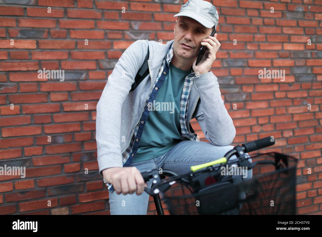 Reifer kaukasischer Mann, der mit dem Telefon spricht, während er auf seinem sitzt Fahrrad in einer Stadt gegen rote Backsteinmauer Stockfoto
