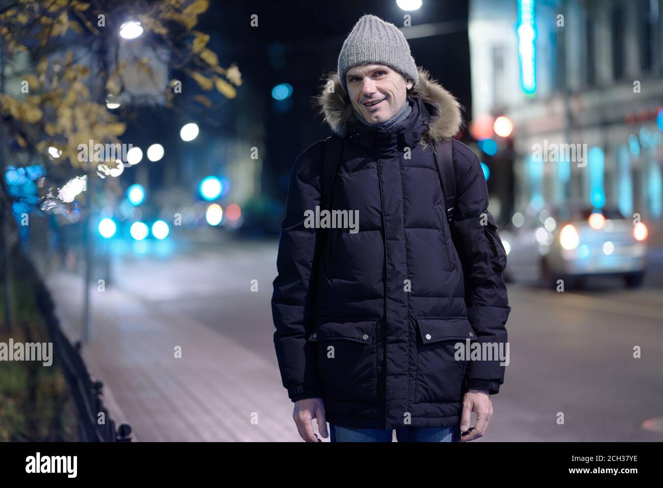Kaukasischer Mann in Winterjacke und Strickmütze steht auf Eine herbstliche Straße einer Nachtstadt und Blick auf Kamera Stockfoto