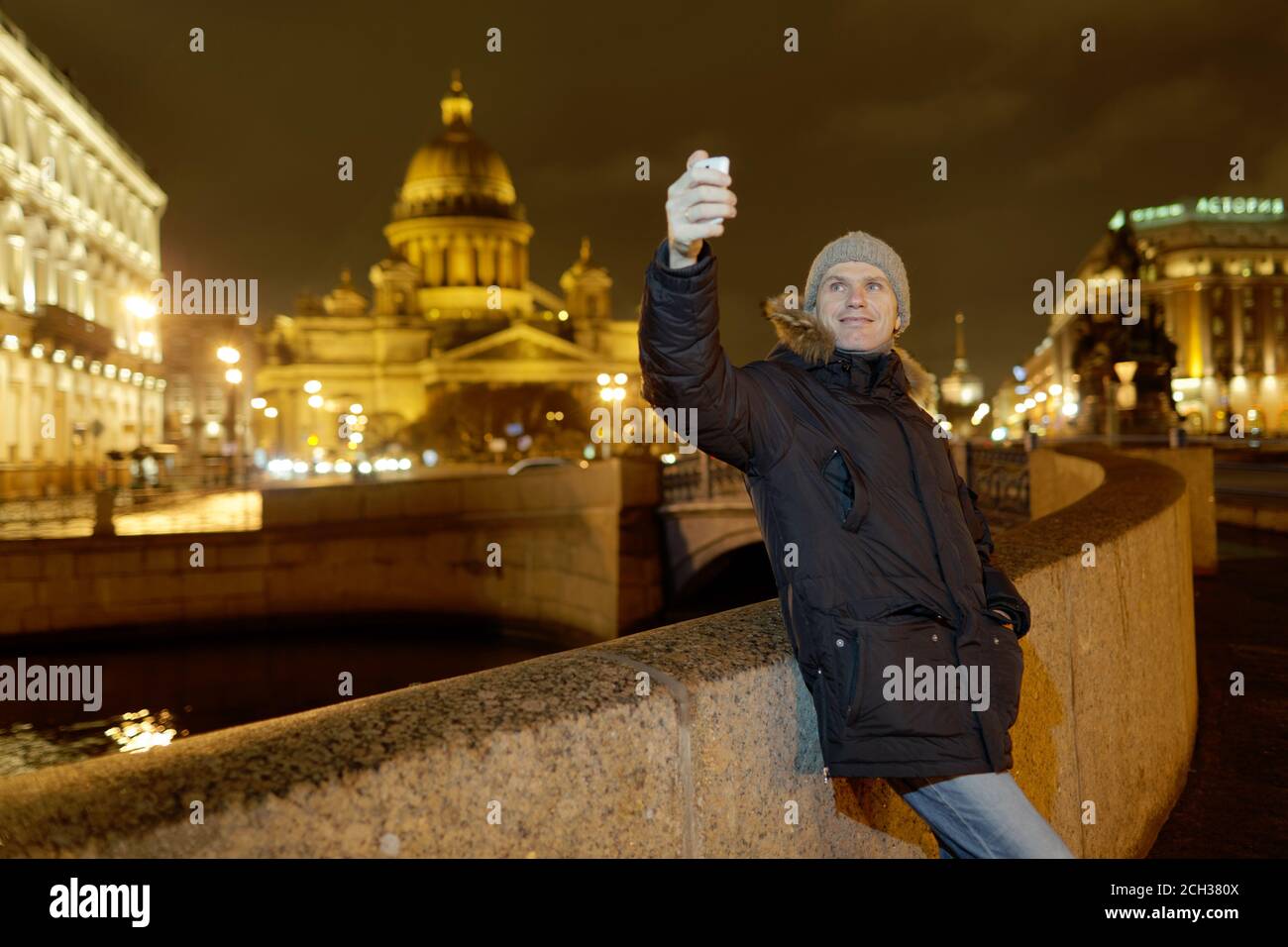 Kaukasischer Mann in Winterjacke macht Selfie mit seinem Smartphone am Ufer des Flusses Moika gegen St. Isaac Kathedrale in Saint-Petersburg, Rus Stockfoto