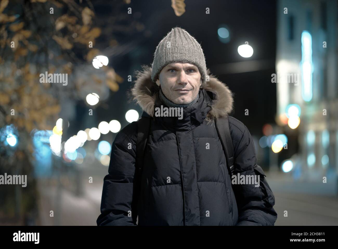 Kaukasischer Mann in Winterjacke und Strickmütze steht auf Eine herbstliche Straße einer Nachtstadt und Blick auf Kamera Stockfoto