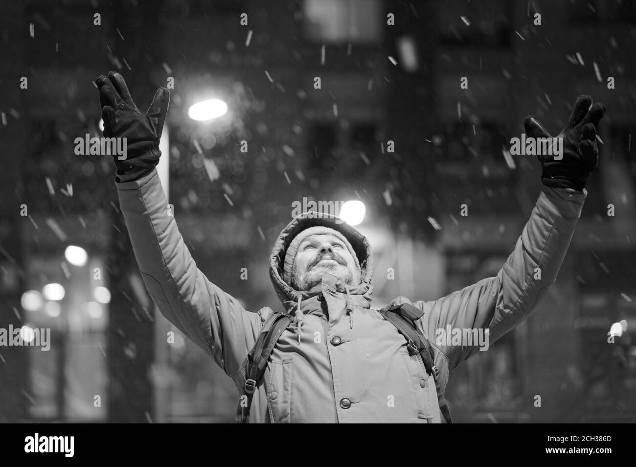 Reifer kaukasischer bärtiger Mann in Winterjacke, der unter Schneefall steht und die Hände zum Himmel erhoben und nach oben schaut. Sepia Stockfoto