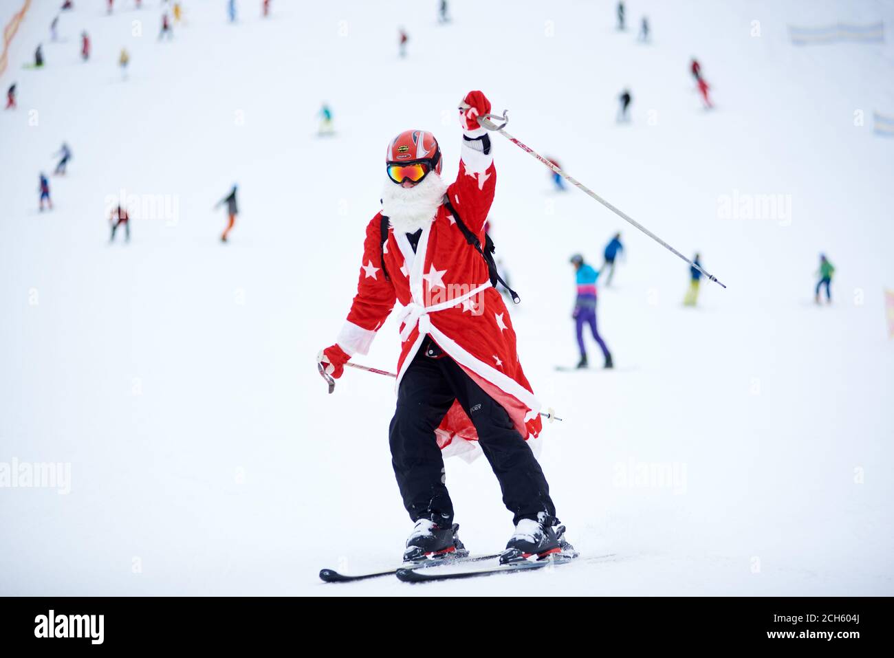 Bukovel, Ukraine - 09. Dezember 2018: Skifahrerin im Weihnachtsmann-Kostüm mit weißem Bart in Schutzbrille und Helm, auf schneebedeckter, hoch gewachsener Piste mit Skistick abwärts und die Hand nach oben Stockfoto