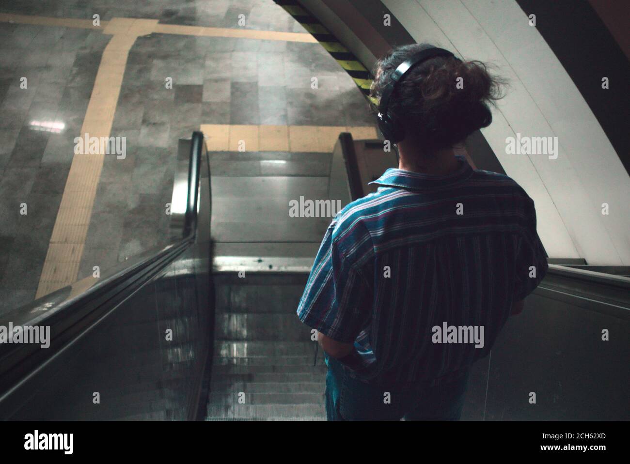 Rückansicht des langhaarigen jungen Mannes, der Musik hört Mit Kopfhörer auf Rolltreppe in der U-Bahn stehen Stockfoto