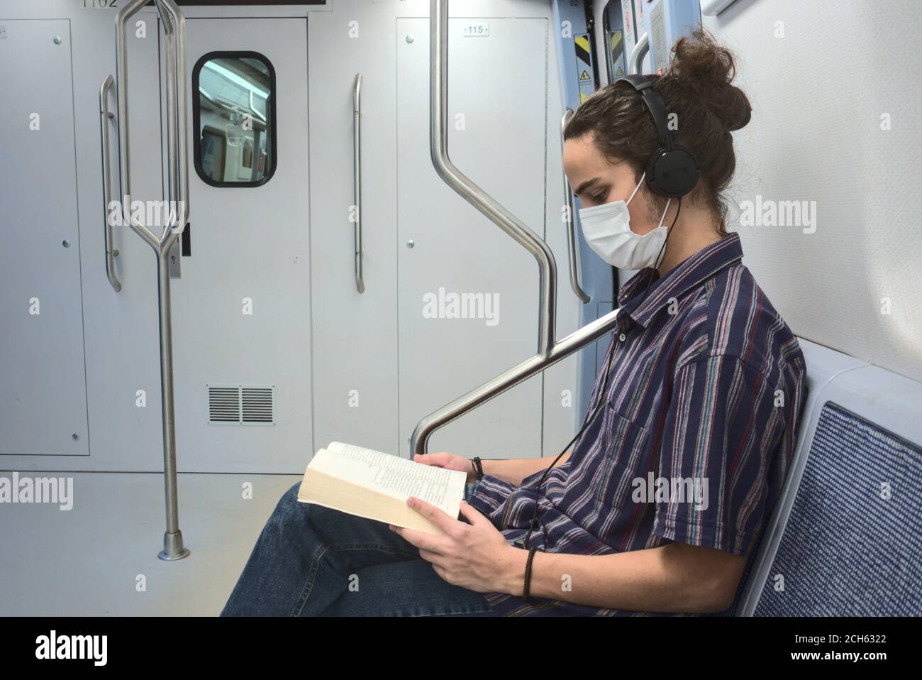 Isoliert und maskiert smart aussehende junge Erwachsene Mann hört zu Musik und Lesen eines Literaturbuches während der Reise in der Blick auf die U-Bahn-Seite Stockfoto