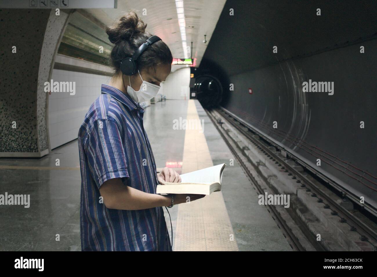 Isoliert maskiert langhaarige junge Erwachsene Musik aus hören Sein Kopfhörer und Literatur Buch lesen, während auf die warten Die U-Bahn kommt Stockfoto