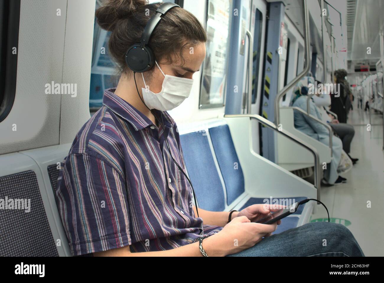 Maskierte langhaarige junge Erwachsene tausendjährige Musik aus hören Sein Kopfhörer während er mit seinem Telefon in der U-Bahn spielte Mit sozialer Distanz p Stockfoto