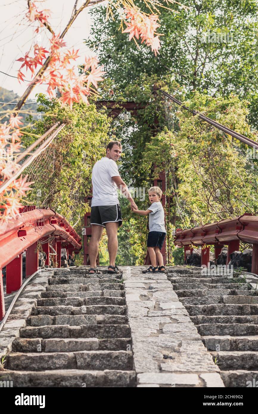 Ansicht von unten Vater Sohn zu Fuß über Steintreppen, die zu roten Eisen Hängebrücke Weg zum Herbst Waldpark. Freundlicher Familienurlaub Stockfoto