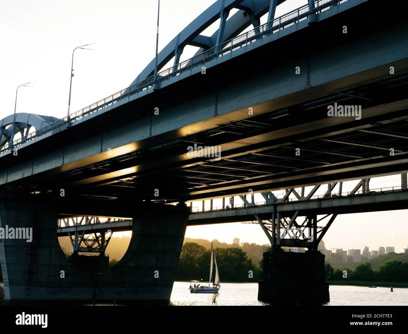 Stadtlandschaft. Eine große Metallbrücke und ein kleines weißes Segelboot im Sommer bei Sonnenuntergang. Stockfoto