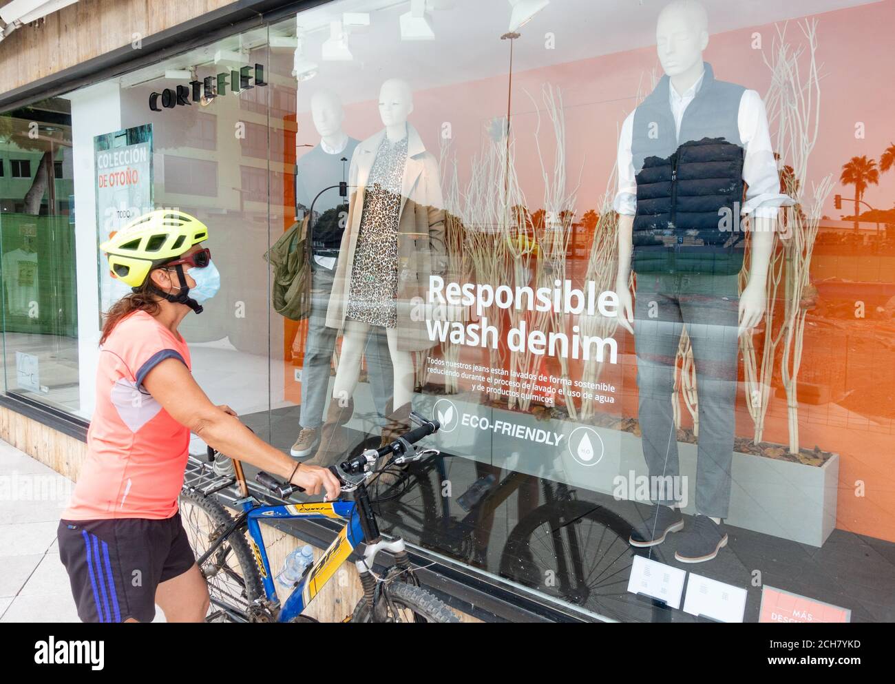 Radfahrerin trägt Gesichtsmaske suchen im Schaufenster mit verantwortungsvollen Wash Denim Aufkleber auf dem Fenster. Schaufensterbummel, umweltfreundliche Kleidung Stockfoto