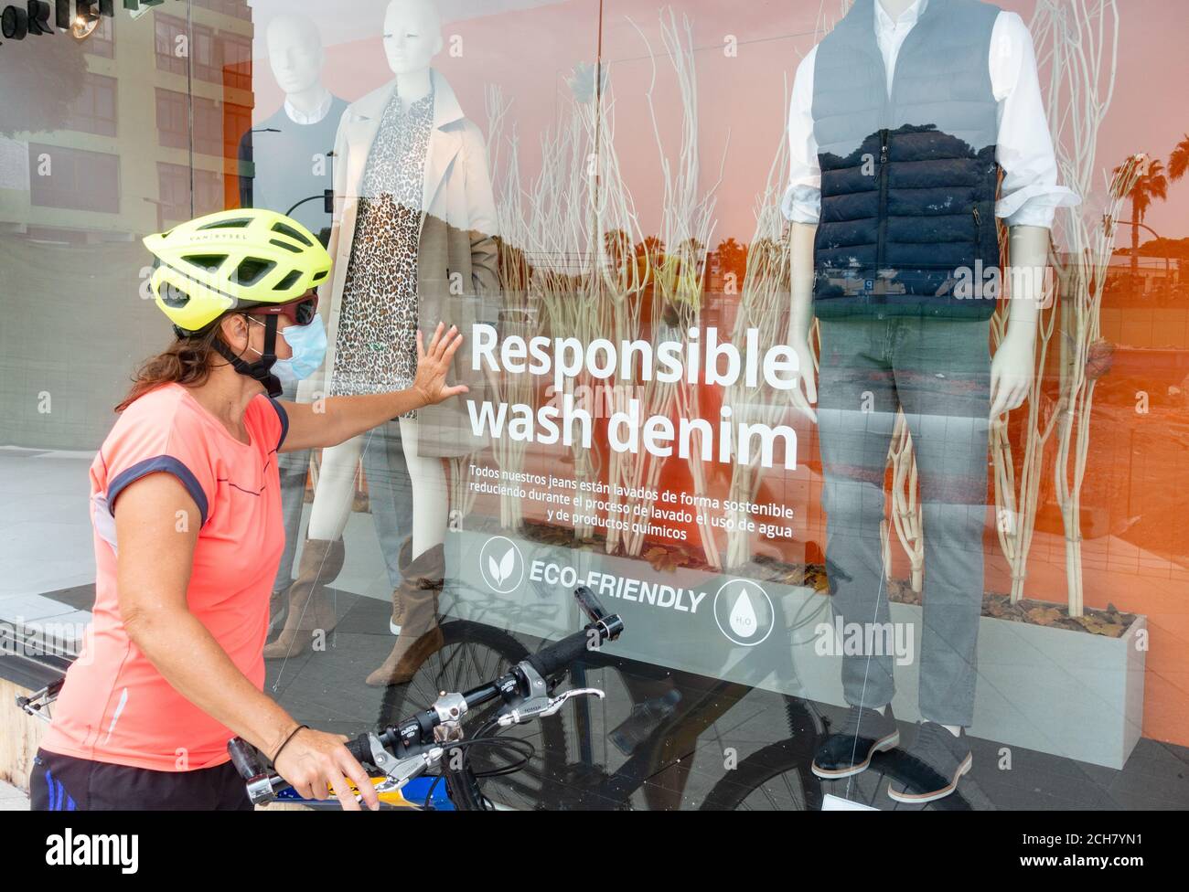 Radfahrerin trägt Gesichtsmaske suchen im Schaufenster mit verantwortungsvollen Wash Denim Aufkleber auf dem Fenster. Schaufensterbummel, umweltfreundliche Kleidung Stockfoto