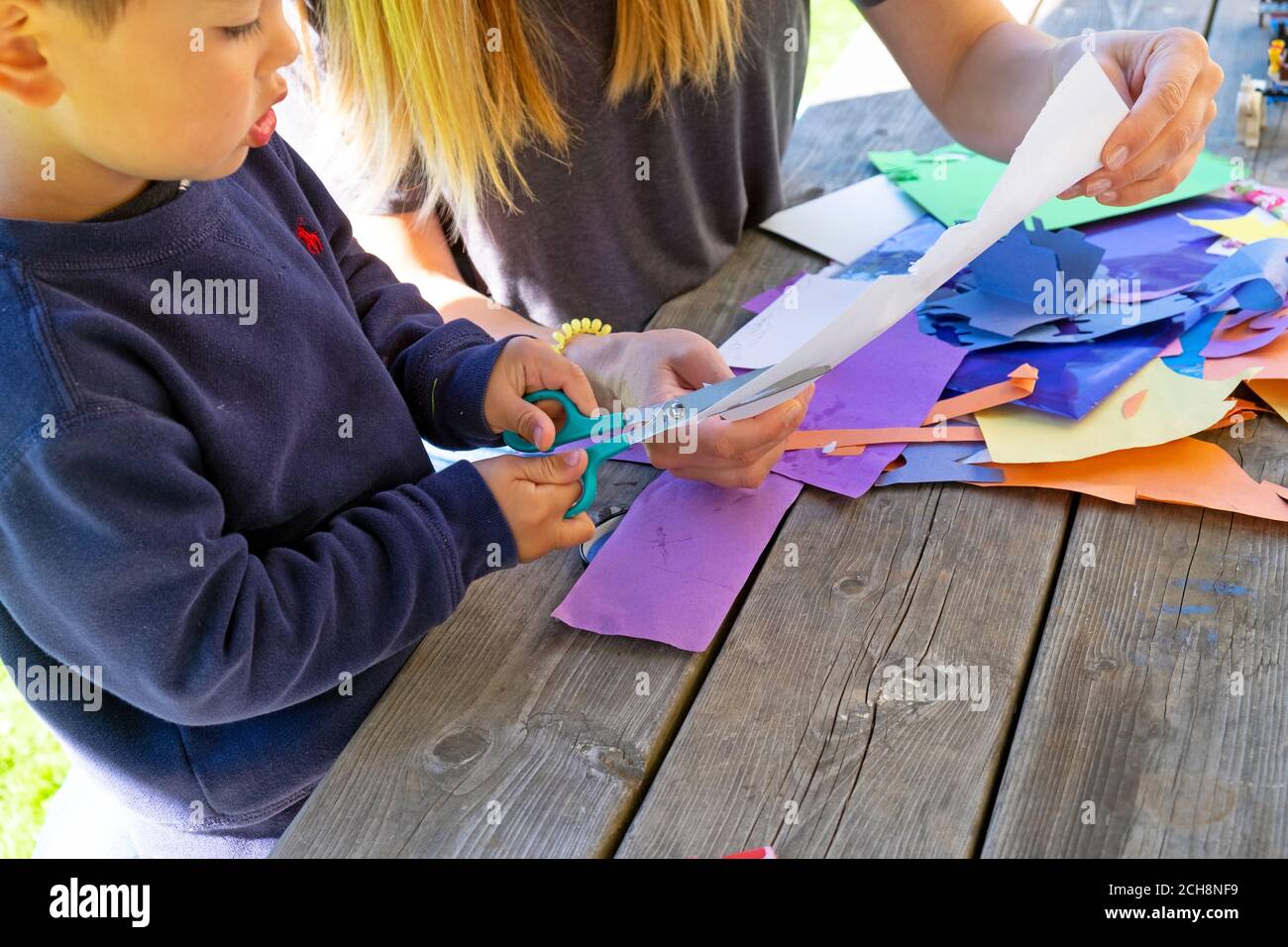 Kleines Kind im Alter von 3 Jahren lernen, Papier mit einer Schere zu schneiden Draußen auf einem Tisch mit buntem Kunstpapier sitzen Carmarthenshire Wales Großbritannien KATHY DEWITT Stockfoto