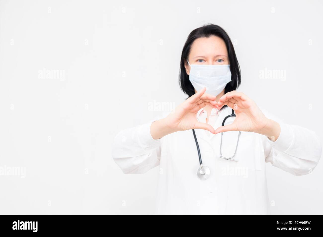 Ärztin in medizinischer Maske zeigt Herz-Symbol auf weißem Hintergrund mit Kopierraum. Stockfoto