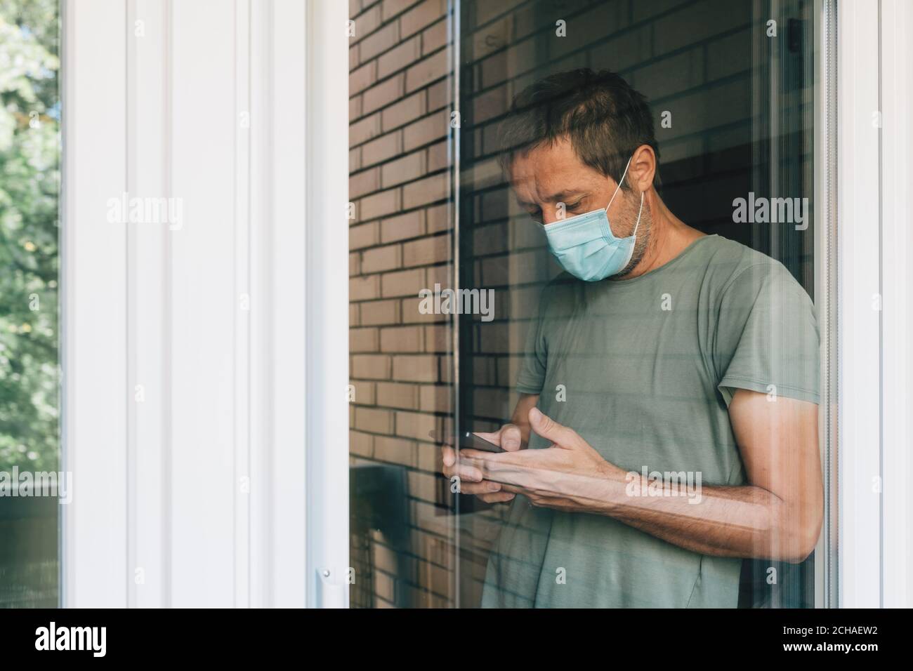 Mann, der während des Coronavirus-Ausbruchs in Selbstisolation auf dem Mobiltelefon SMS schreibt, Porträt eines erwachsenen Mannes hinter dem Fenster Stockfoto