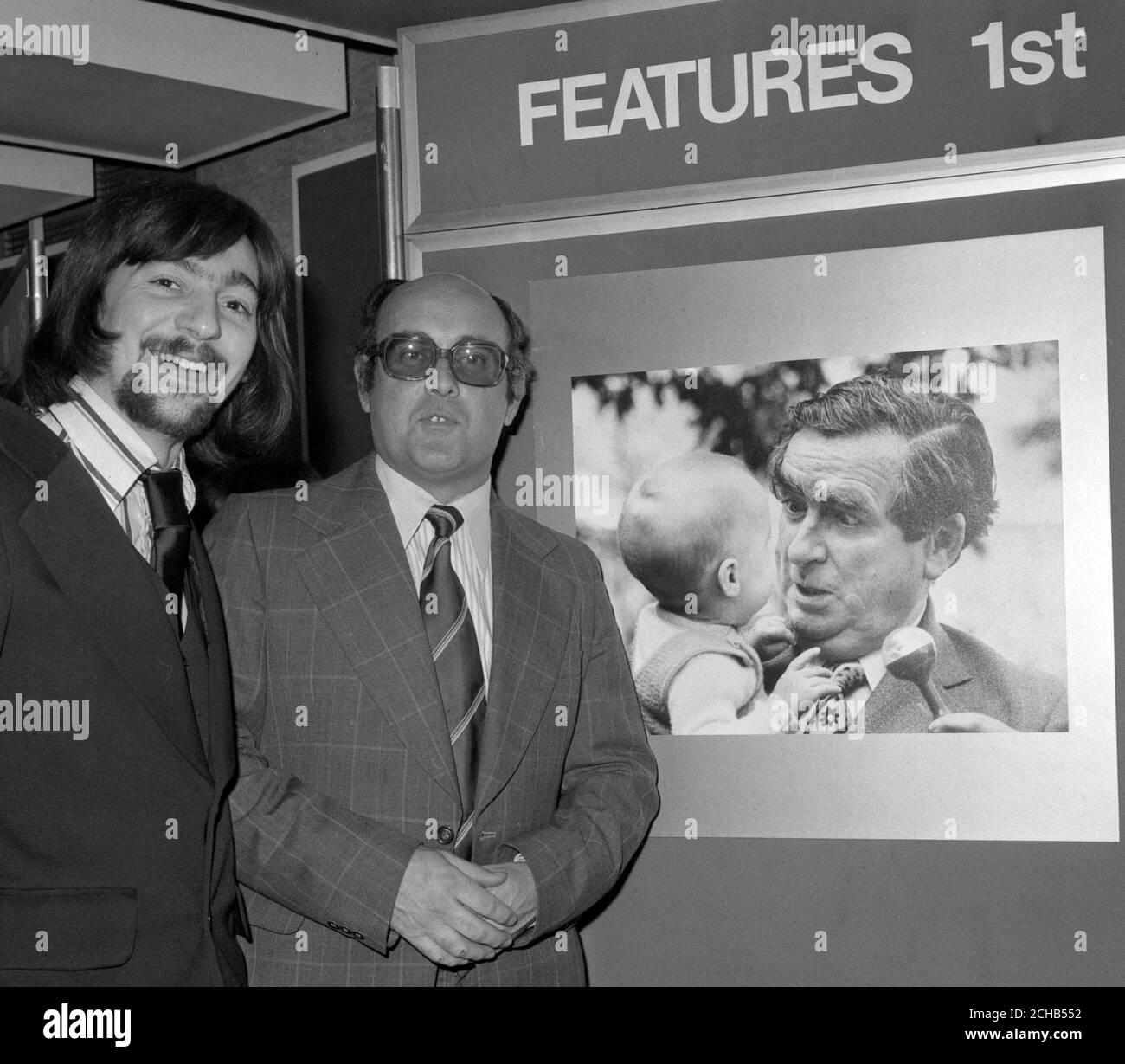 Bei der £1,000 Ilford Print Awards Zeremonie der Präsentationen im Sudbury House, Newgate Street, London, stehen neben dem Gewinnerbild der Mitarbeiter der Press Association, Stanley 'Jimmie' James (r), der den ersten Preis in der Rubrik Features gewann, und der Fotodrucker der Press Association, Derek 'Pip' Piper. Es zeigt Denis Healey, den Schatzkanzler, mit einem Baby. Stockfoto