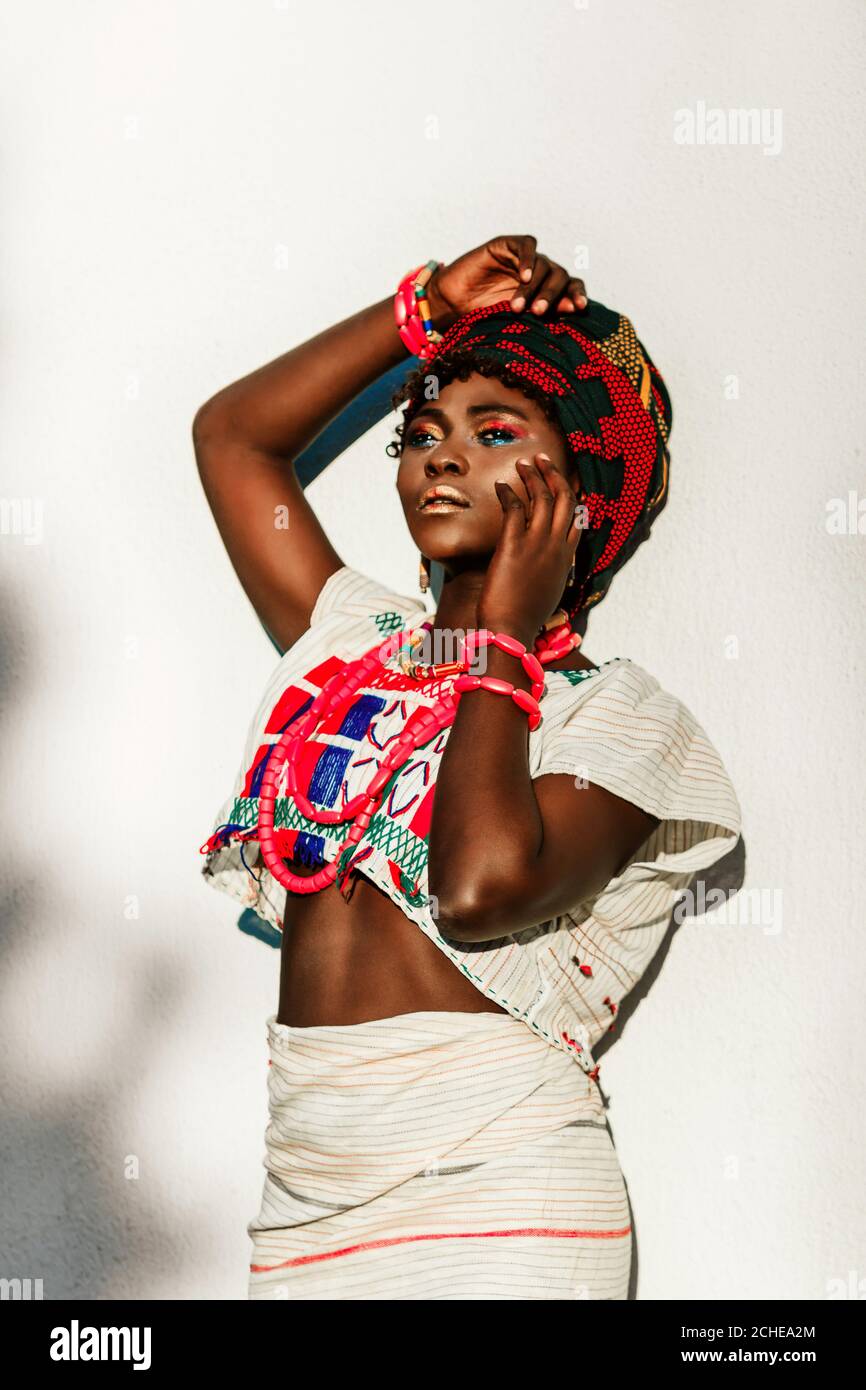 Zugeschnittenes Bild von afrikanischen weiblichen Modelin Nationalkostüm, Schmuck, Turban und Make-up posiert in über weißen Wand Hintergrund, Tageslicht Sonne. Stockfoto