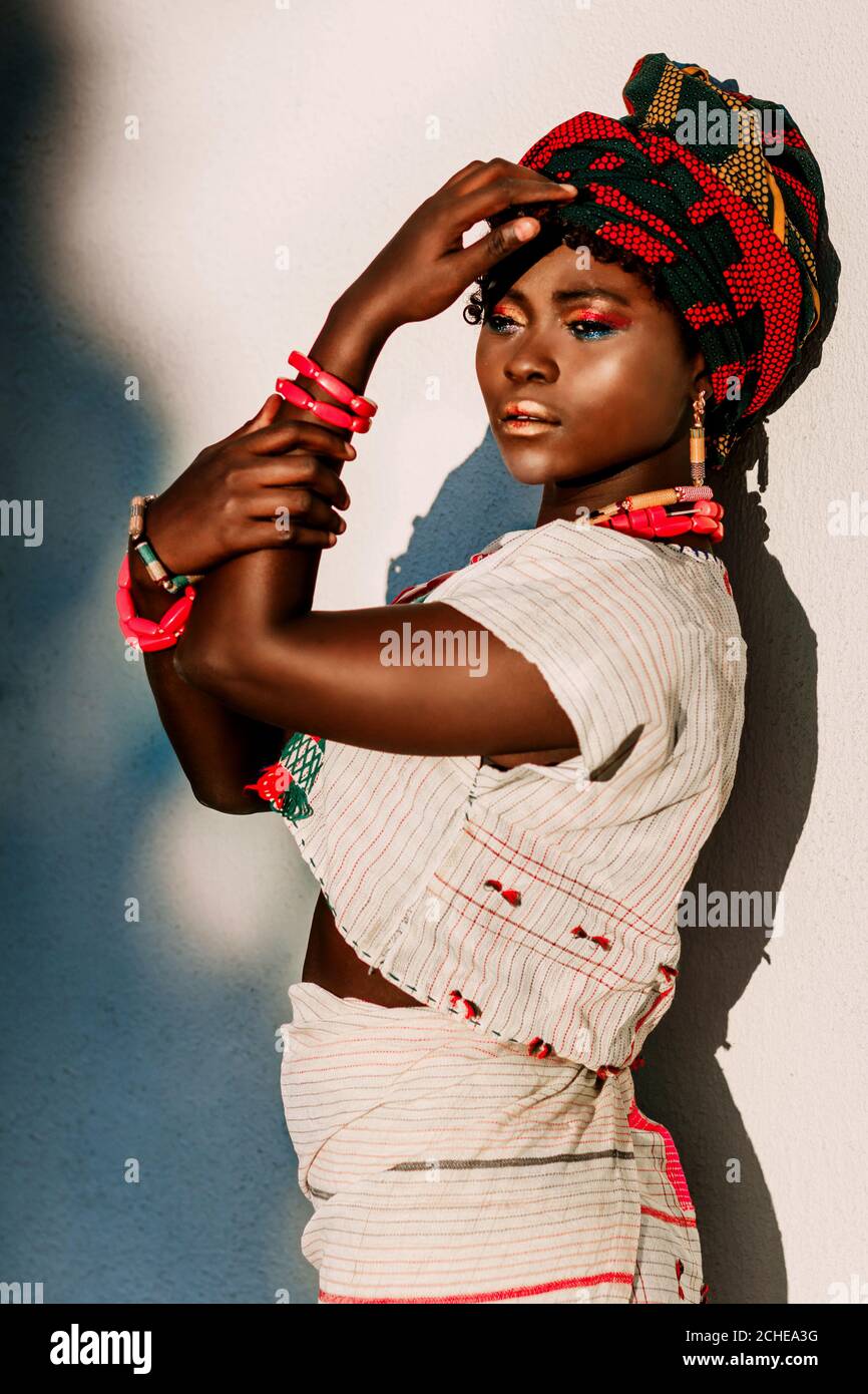 Zugeschnittenes Bild von afrikanischen weiblichen Modelin Nationalkostüm, Schmuck, Turban und Make-up posiert in über weißen Wand Hintergrund, Tageslicht Sonne. Stockfoto