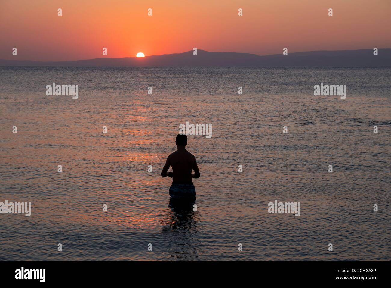 Orangefarbene Sonne untergeht im Morgengrauen hinter den Bergen. Schwarze Silhouette eines jungen Mannes, der in farbenfroh blauem Wasser steht und von kleinen Wellen umgeben ist Stockfoto