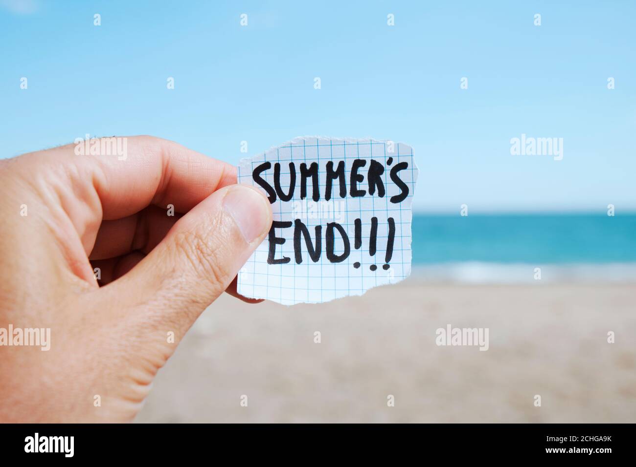 Nahaufnahme der Hand eines jungen kaukasischen Mannes am Strand, vor dem Ozean, hält ein Stück Papier mit dem Text Sommer Ende in ihm geschrieben Stockfoto