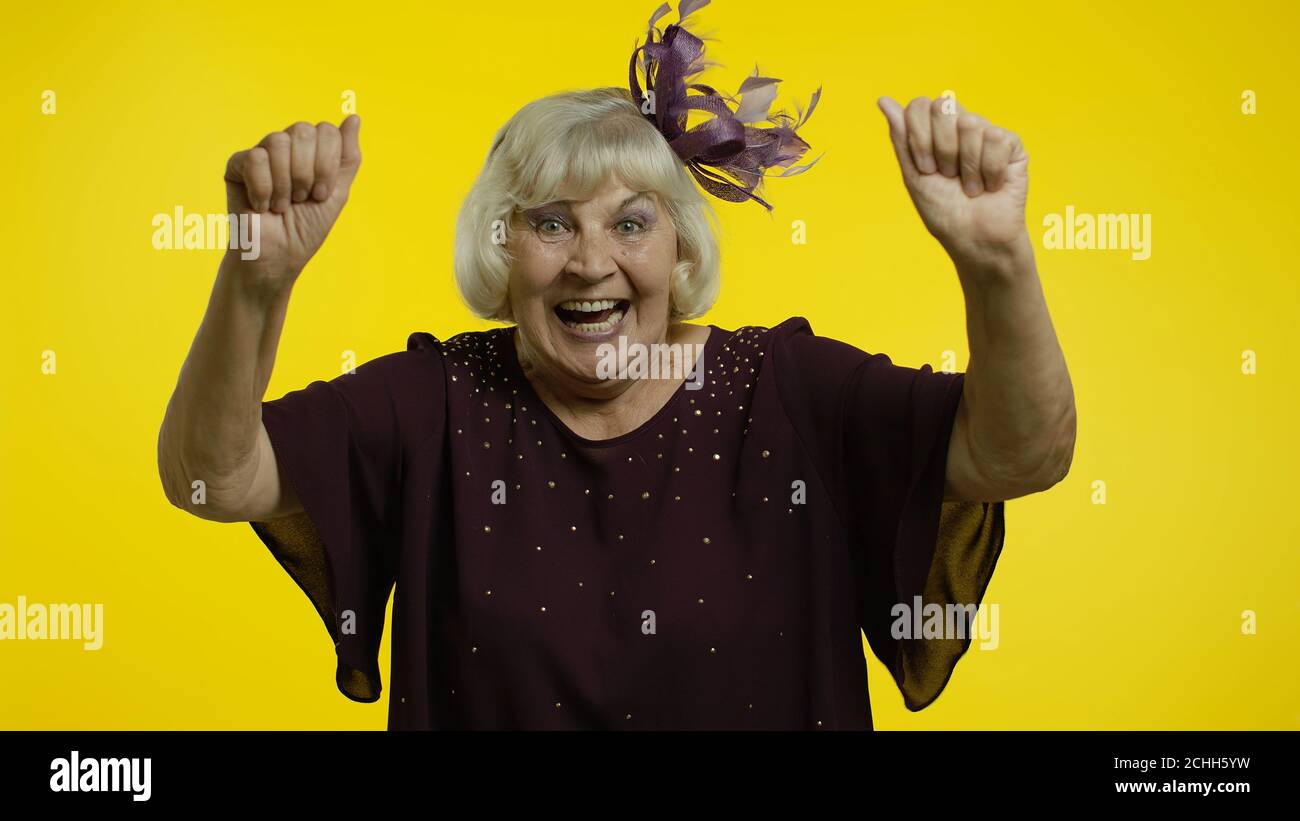 Extrem glücklich zufrieden ältere Frau mit blonden Haaren feiert Sieg mit  toothy Lächeln, Erfolg. Freude an der Gewinnerwertung. Ältere stilvolle  Dame Oma auf gelbem Hintergrund Stockfotografie