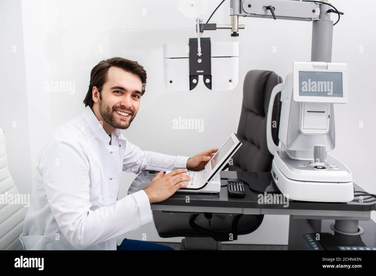 Lächelnder Optiker sitzt auf seinem Arbeitsplatz in einer modernen Klinik. Augenuntersuchung und Sehdiagnostik Stockfoto