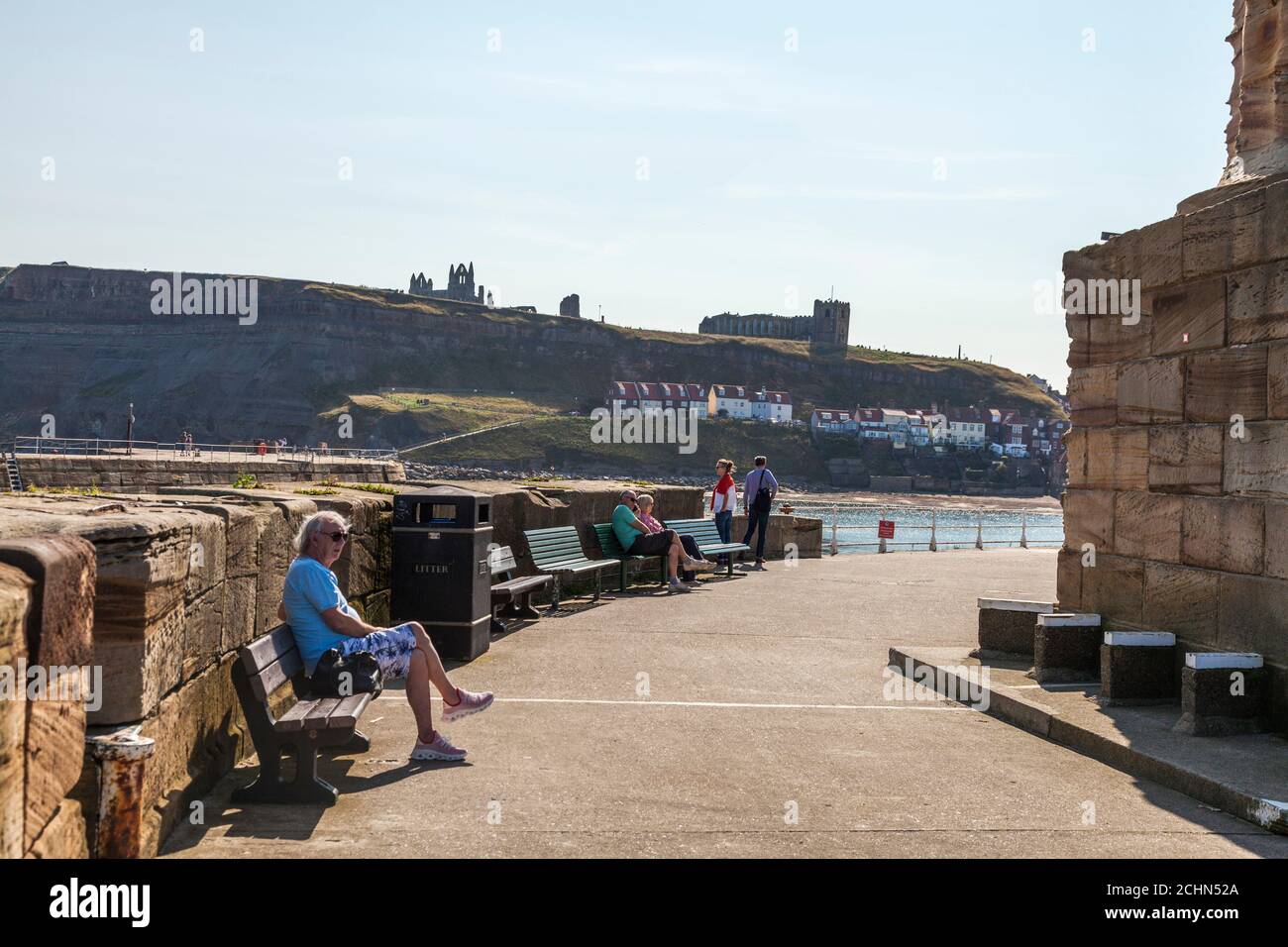 Die Leute saßen entspannt auf Bänken auf dem Pier in Whitby, North Yorkshire, England, Großbritannien mit der Abtei und der Kirche im Hintergrund Stockfoto