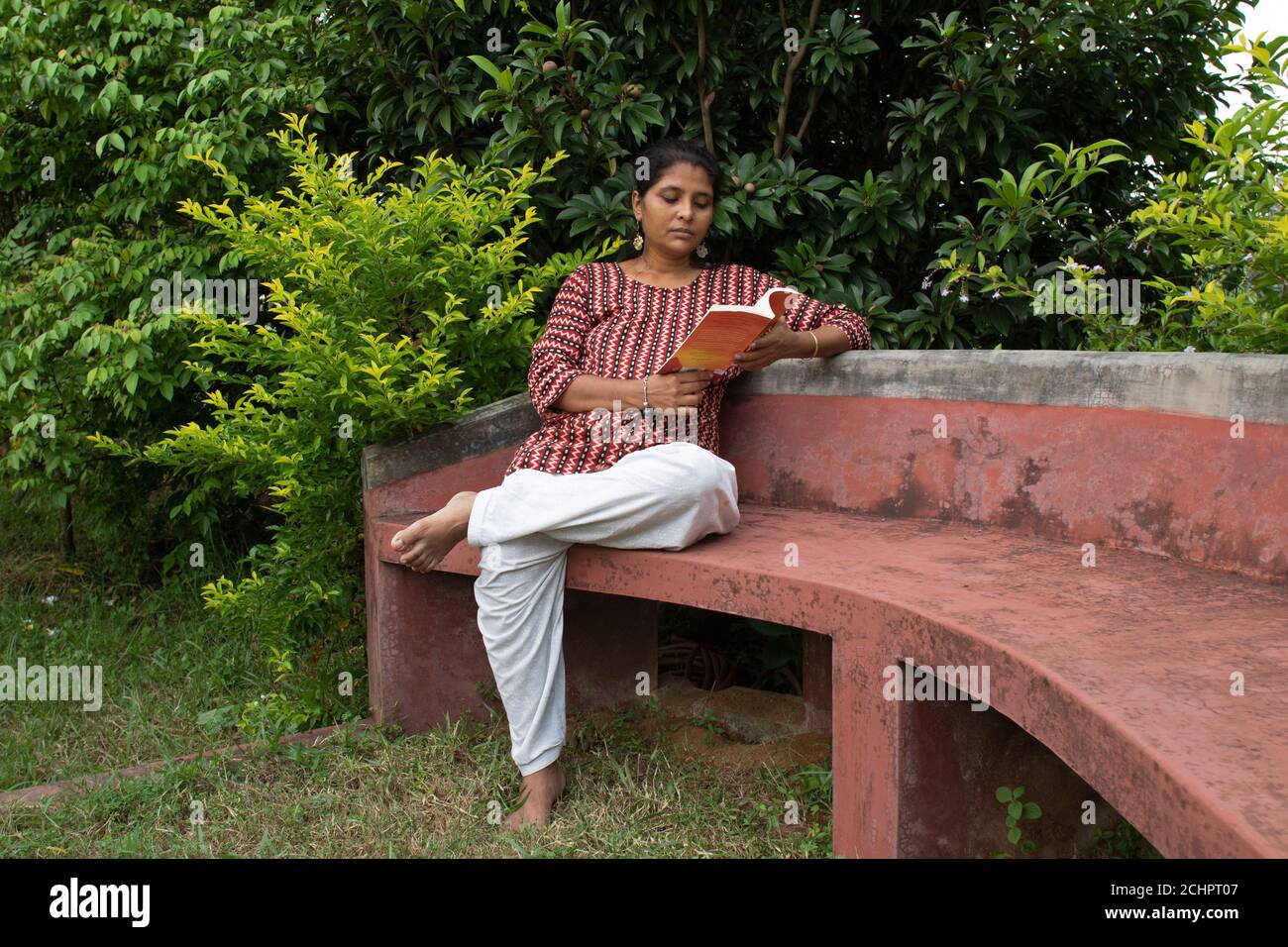 Eine schöne Frau in legerer Kleidung, die auf einer roten Bank sitzt, liest ein Buch und denkt über die Geschichte in einem Park nach Stockfoto