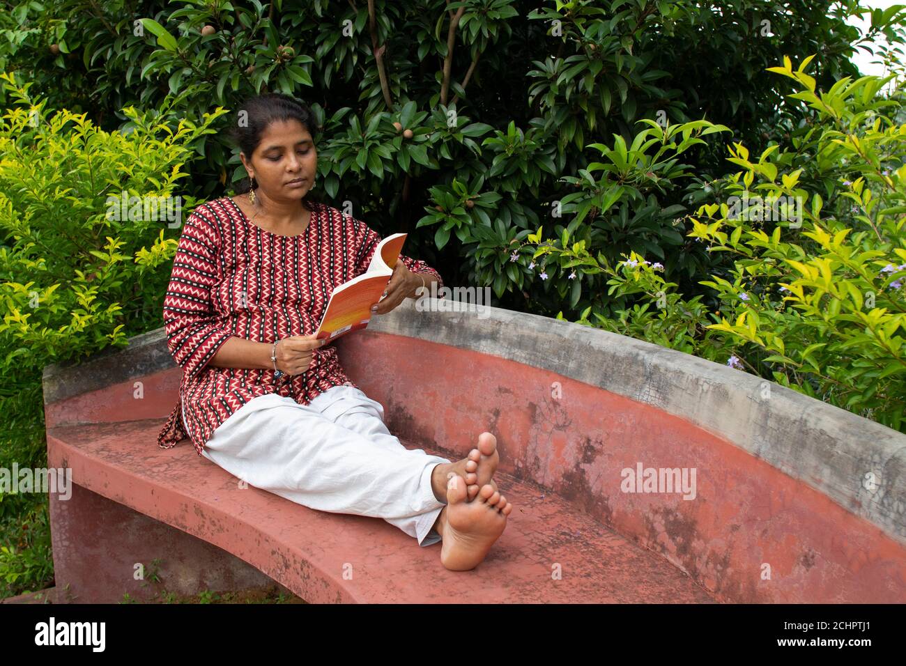 Eine schöne Frau in legerer Kleidung, die auf einer roten Bank sitzt, liest ein Buch und denkt über die Geschichte in einem Park nach Stockfoto