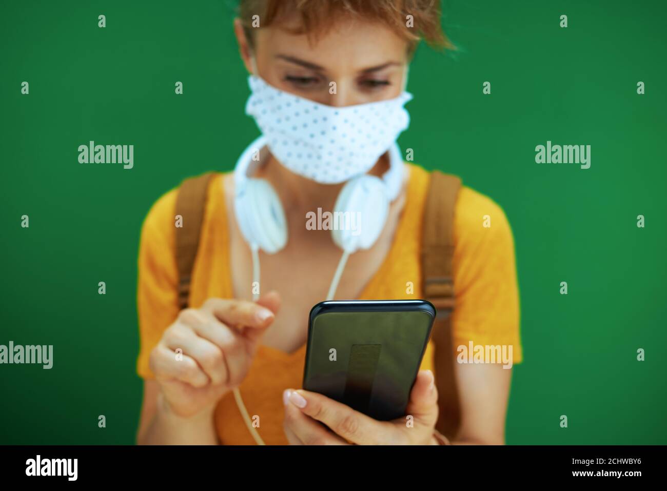 Leben während covid-19 Pandemie. Student in gelbem Hemd mit medizinischer Maske, weißen Kopfhörern und Rucksack senden Textnachricht von Smartphone isoliert Stockfoto