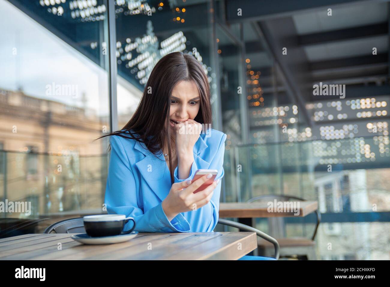 Beißende Faust beim Blick auf Handy. Nahaufnahme Porträt einer Frau in blauen Business-Anzug wütend mit ihrem Handy in der Café-Terrasse Stockfoto