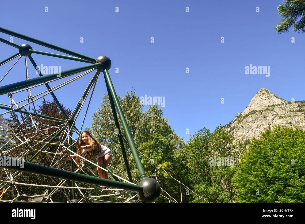 Ein kleines Mädchen (10 J.o.), das im Sommer auf einem Spielplatz mit dem Mont Chetif im Hintergrund auf einer Spielanlage klettert, Courmayeur, Aosta, Italien Stockfoto